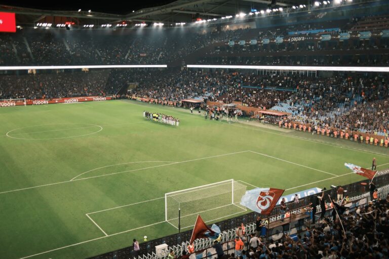 Stadion de fotbal luminat seara, cu tribunele pline de spectatori în timpul unui meci